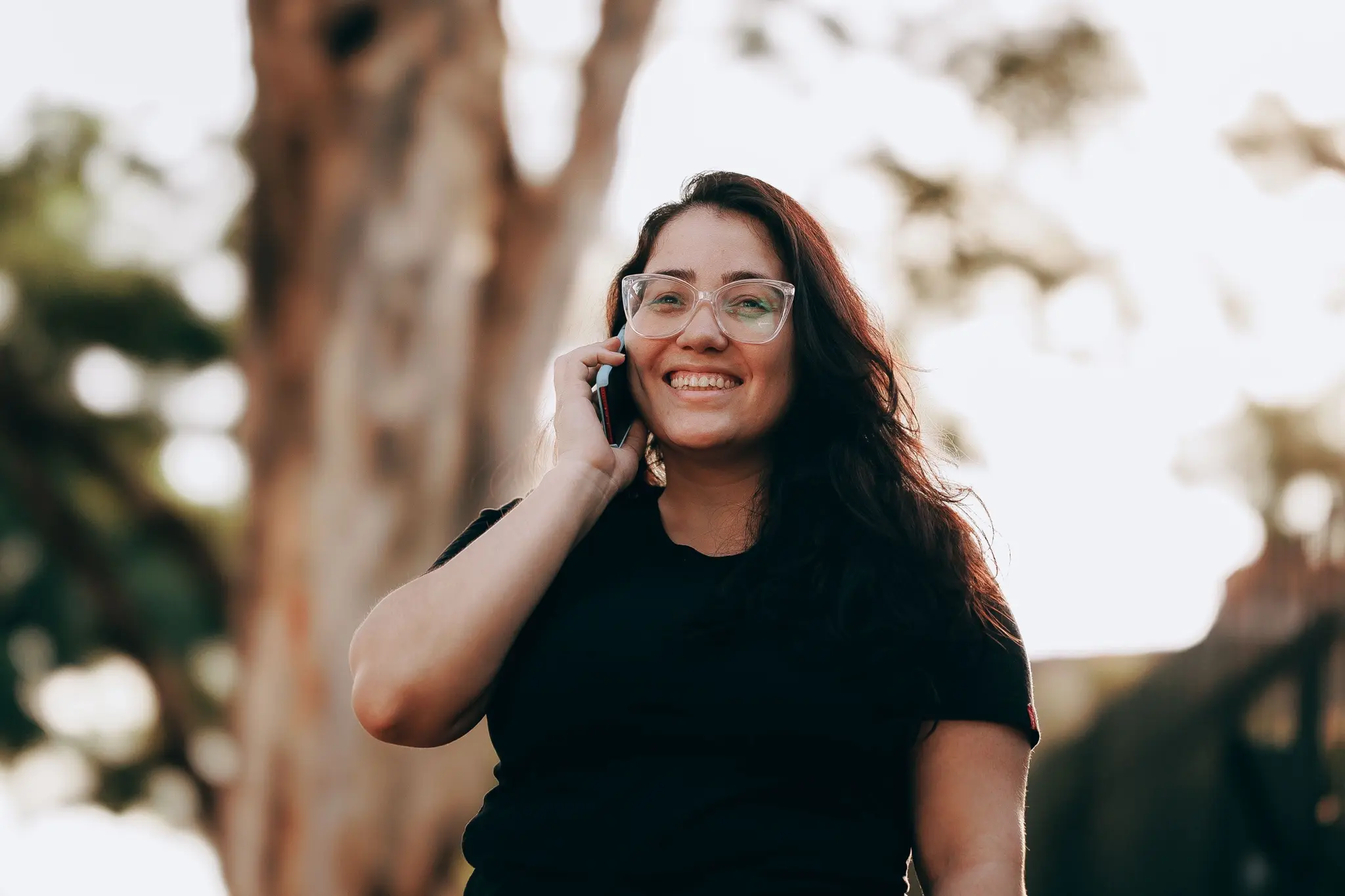 Una mujer de lentes ríe mientras habla por teléfono bajo los árboles